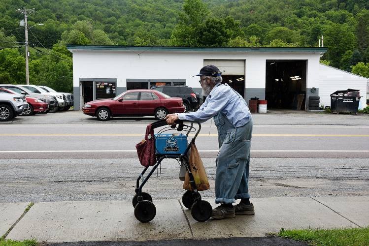 Elderly person using a walker on a sidewalk near a road, with a red car and a building in the background. Trees and greenery are visible behind the building.