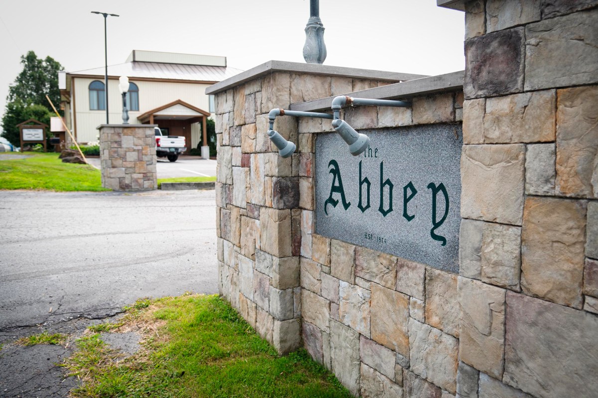 Stone sign with "The Abbey" engraved, mounted on a stone wall at the entrance to a property with buildings in the background.
