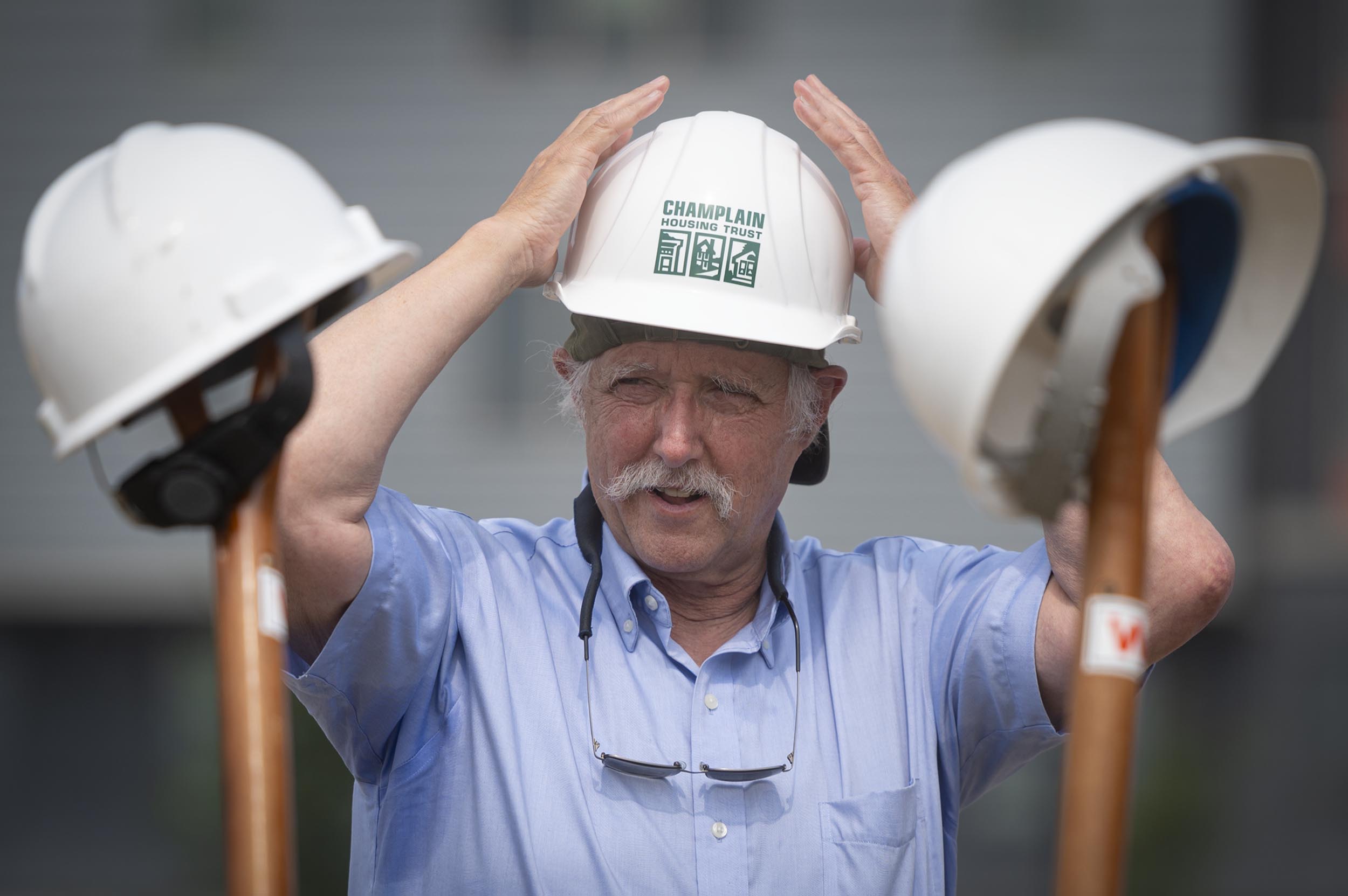 An older man in a blue shirt adjusts a hard hat labeled "CHAMPLAIN," with two other hard hats resting on shovels in the foreground.