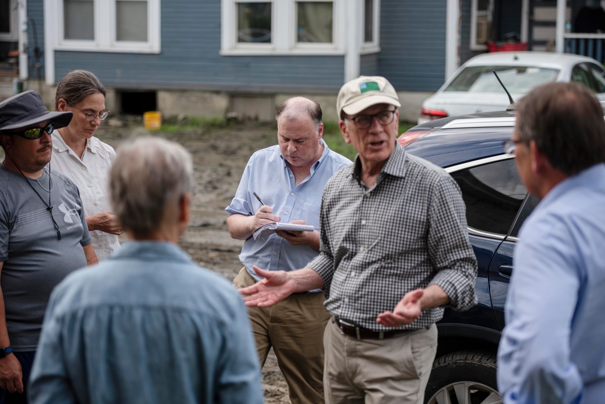 A group of five people stand outdoors near a blue house. One man is speaking while others listen and take notes. A car is parked nearby.