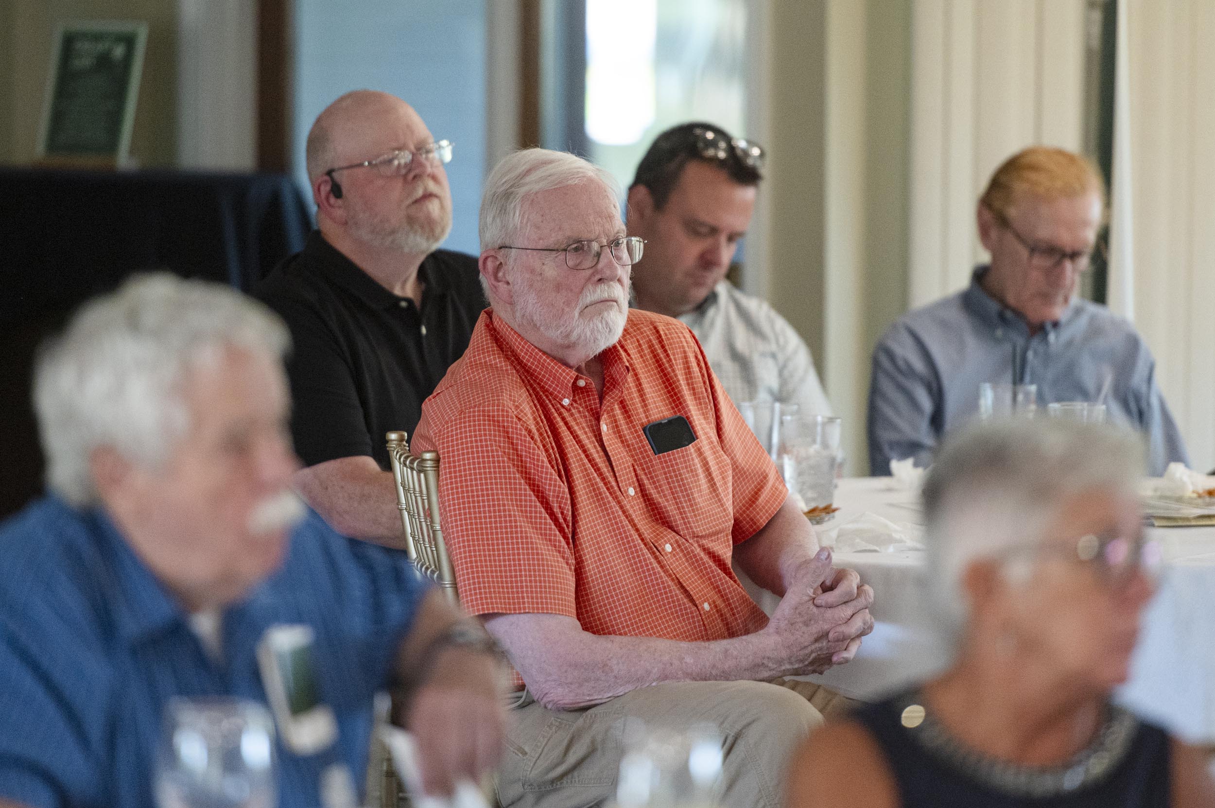 A group of older adults sits at tables, attentively listening during an indoor event.