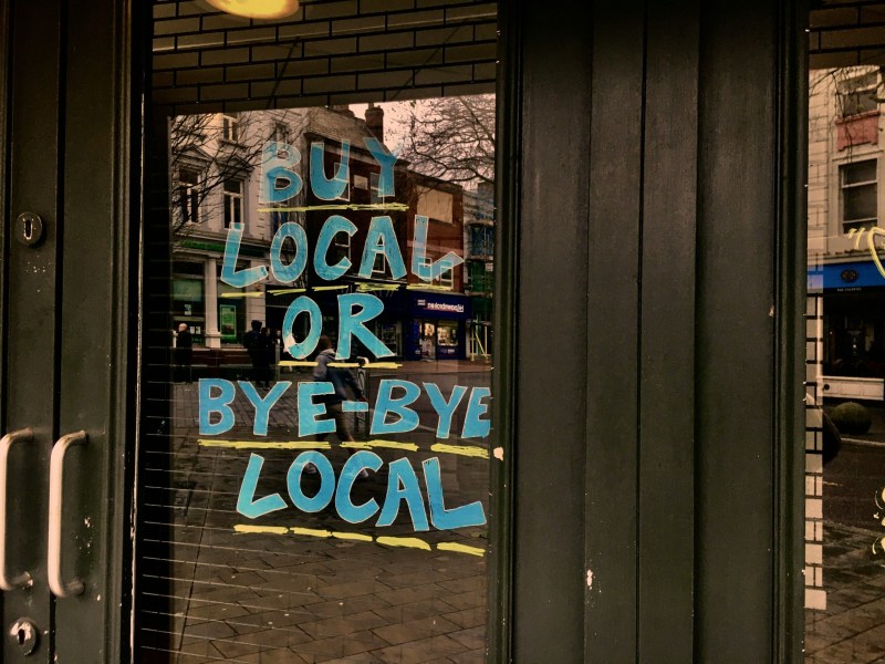 Glass door with the handwritten message: "Buy local or bye-bye local" in blue letters. Reflections of buildings and street visible.