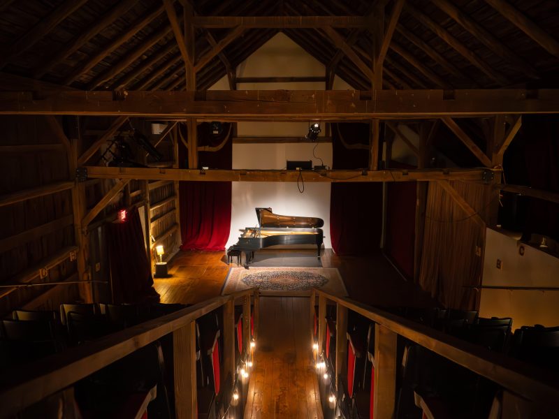 A grand piano on a dimly-lit stage inside a rustic, wooden theater with rows of seats on either side of a central aisle.