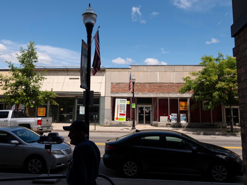 A street scene showing parked cars, a streetlamp with flags, and storefronts with “For Lease” and “For Rent” signs on a sunny day.