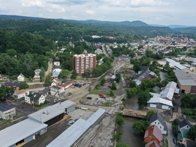 Aerial view of a small town with a mix of residential and industrial buildings, muddy roads, and visible flooding along a riverbank.