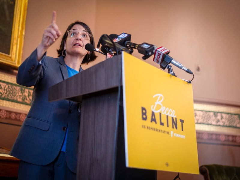 Person in a blue suit speaks passionately at a podium with multiple microphones and a sign that reads, "Becca Balint, US Representative, Vermont.