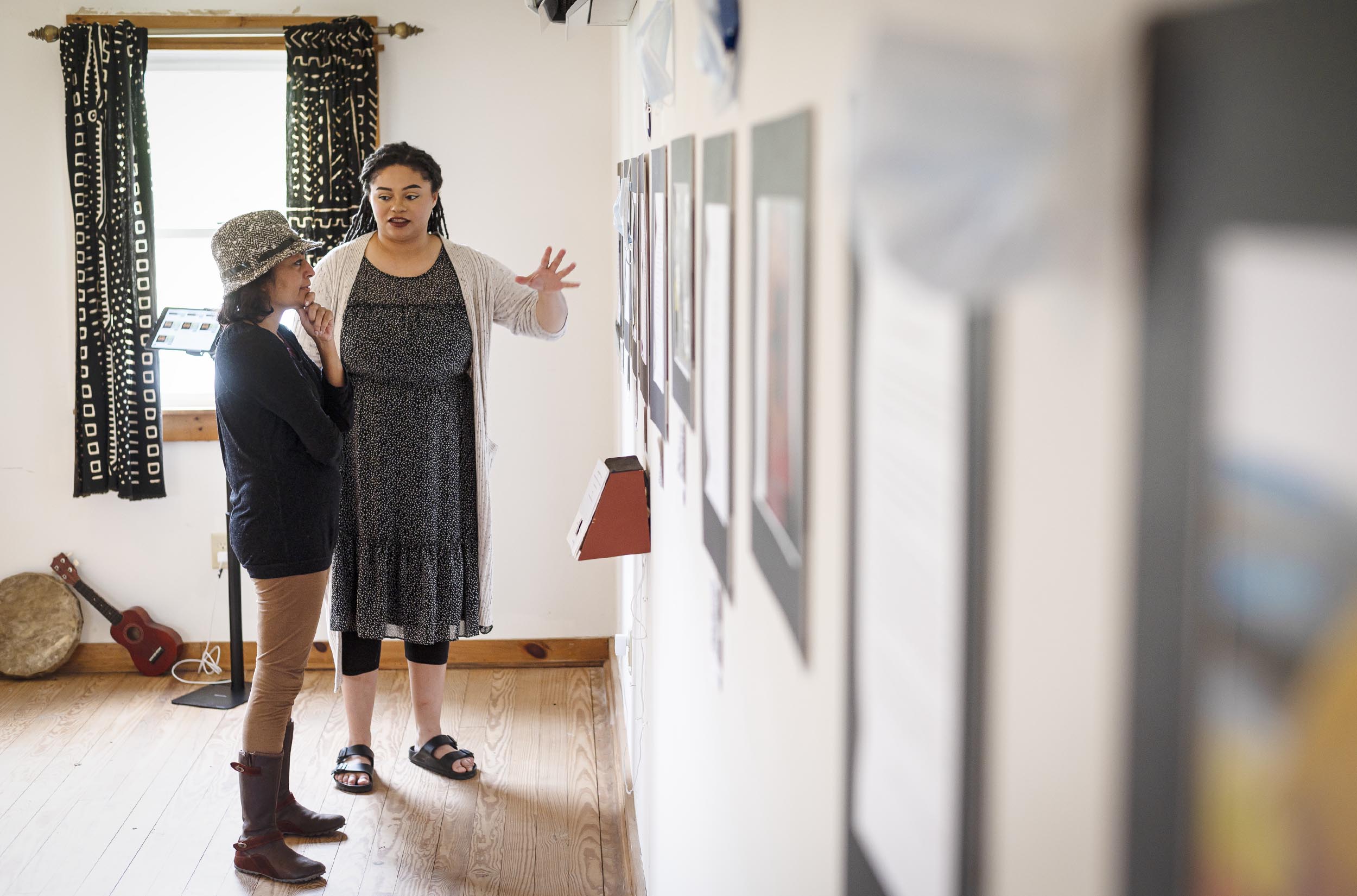 Two women discussing an art exhibit in a gallery with white walls adorned with various framed artworks.
