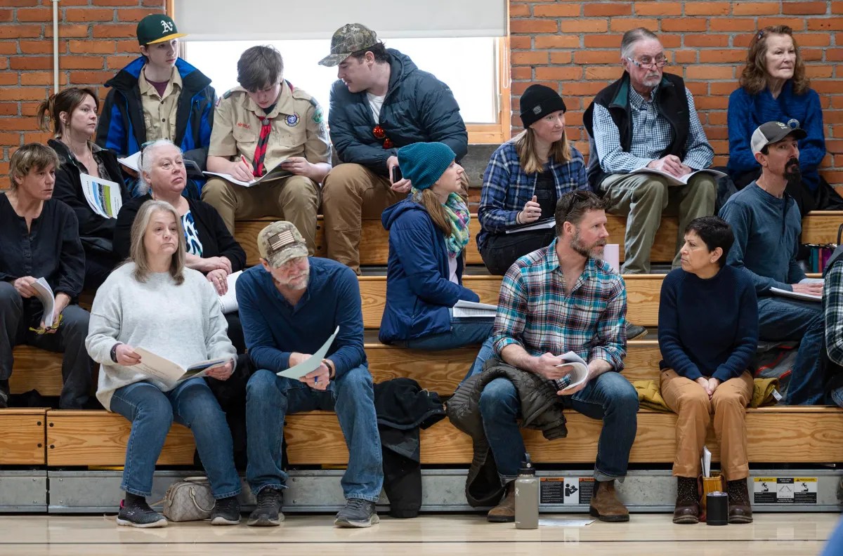 People sitting on bleachers in a brick-walled room, some holding papers. They appear to be listening or waiting, dressed in casual clothing, including hats and jackets.
