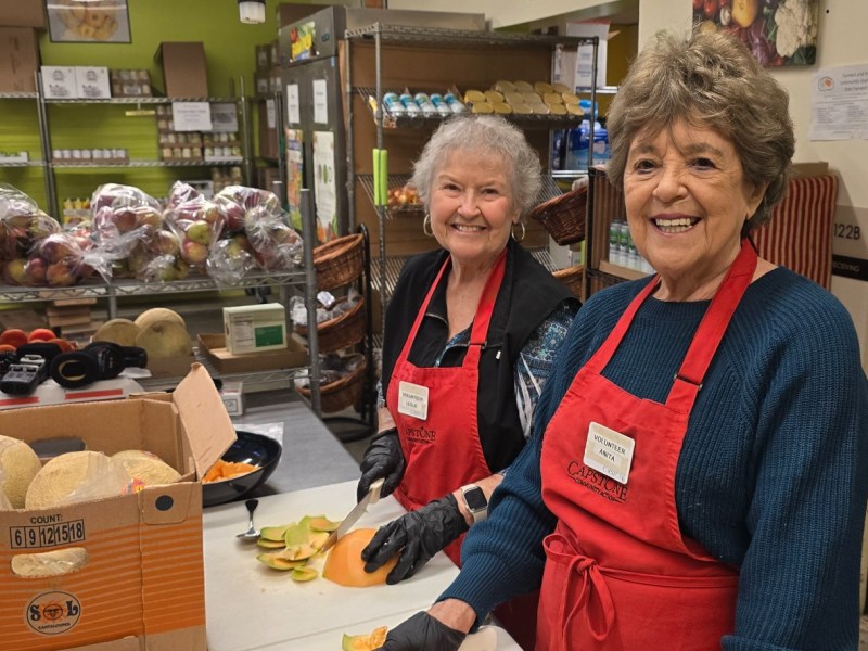 Two women wearing red aprons and gloves prepare food at a kitchen counter, with fresh produce and pantry shelves visible in the background.