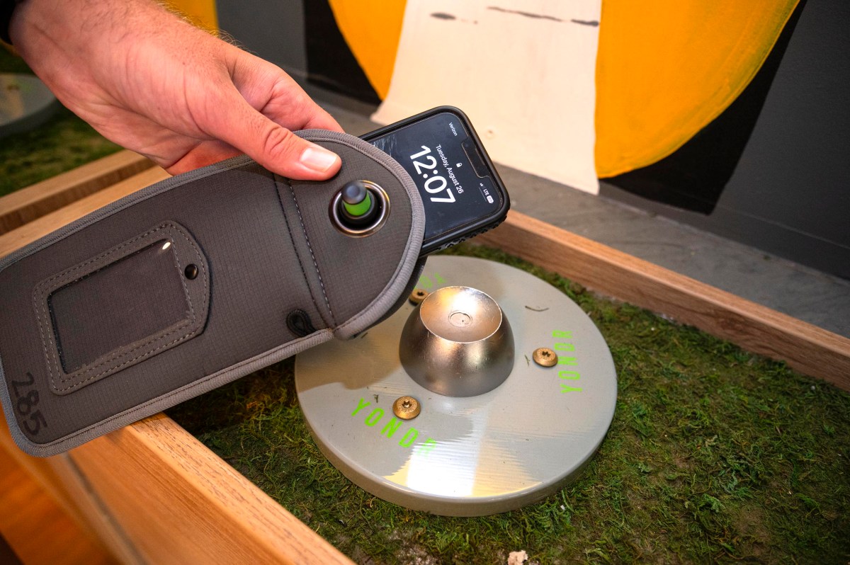 A person scans a round metal object labeled "YOND" with a smartphone inside a grey pouch, placed on a grassy surface in a wooden enclosure.