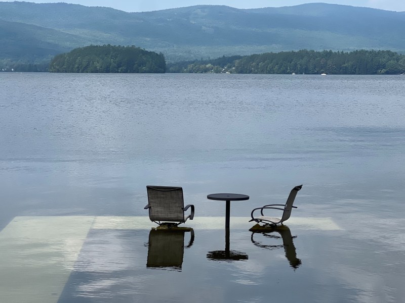 Two empty chairs and a small table partially submerged in water, with a scenic view of a lake and forested mountains in the background.