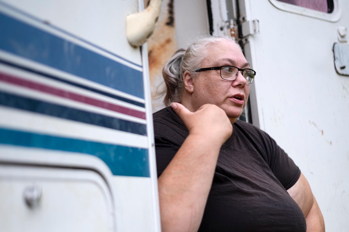 A woman with glasses and gray hair pulled back is talking while standing outside next to a white vehicle with blue and red stripes.