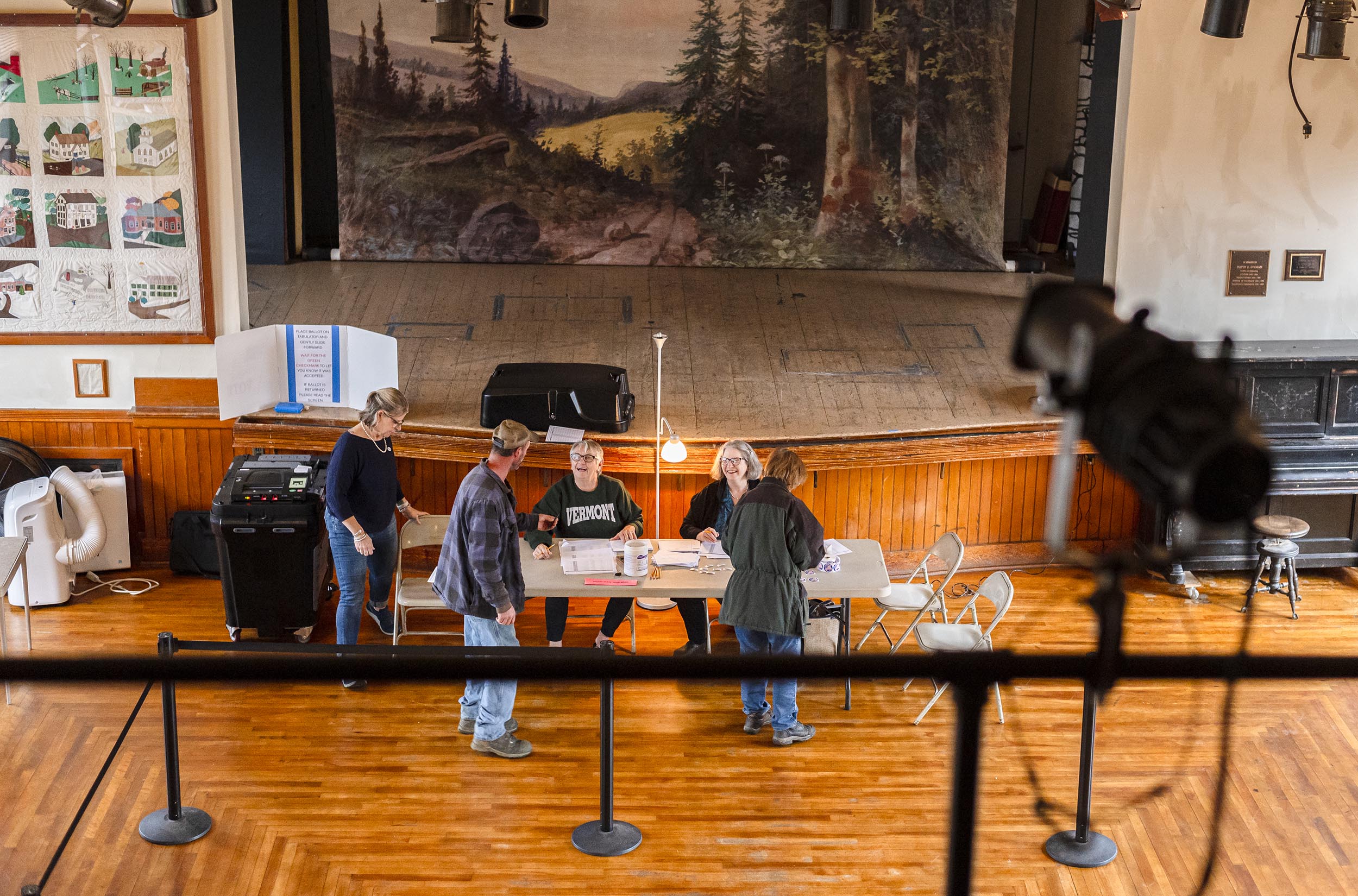 People are gathered around a table in a hall with a stage in the background. A spotlight is facing the stage, and quilts are displayed on the wall.