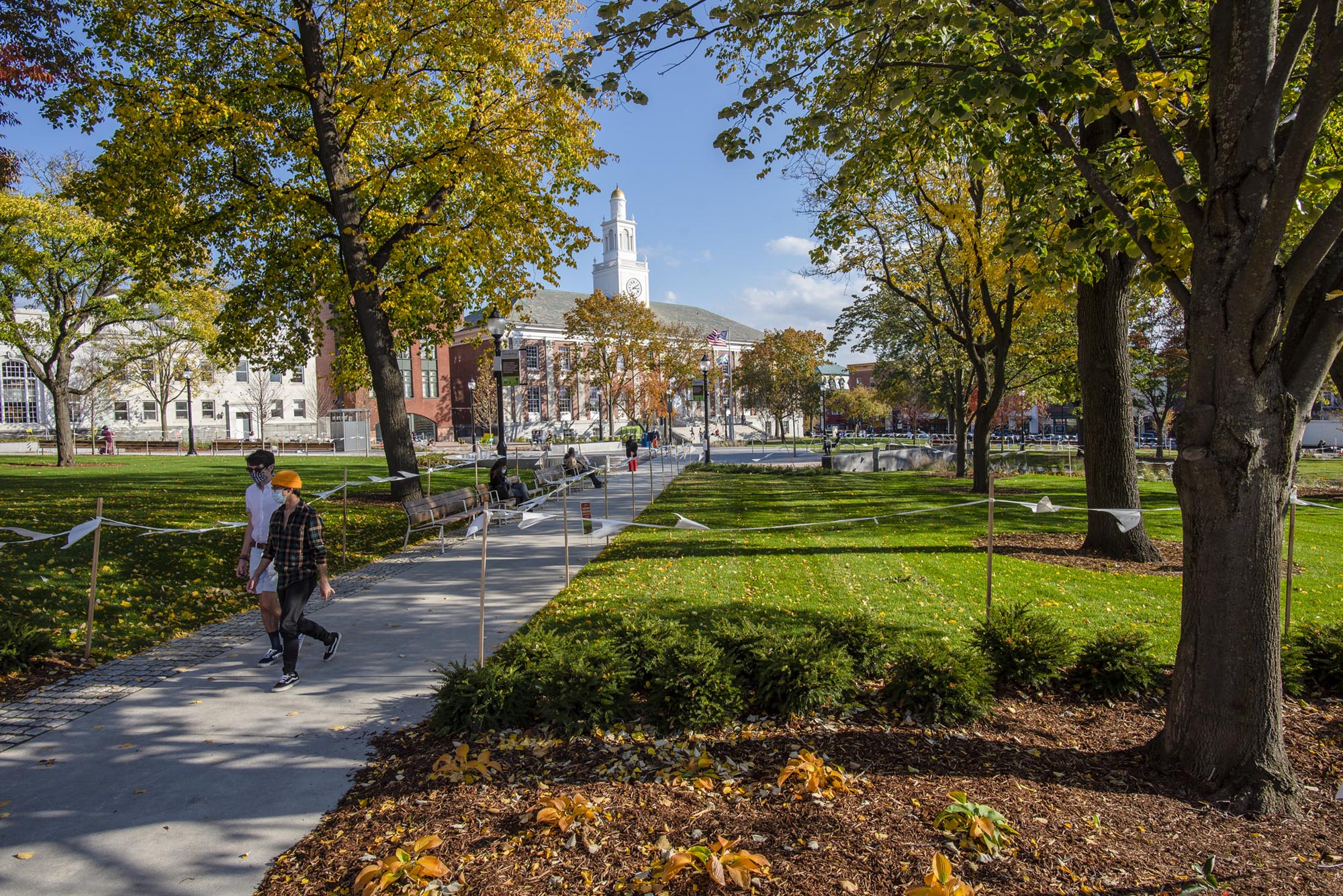 People walk along a paved path through a landscaped campus lawn with trees and a large academic building in the background on a sunny day.