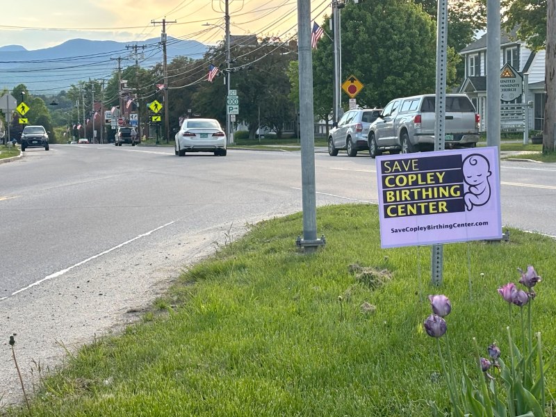A "Save Copley Birthing Center" sign is displayed on a grassy roadside, with cars parked along the street and mountains visible in the background.