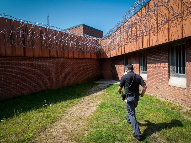 A man in a uniform walks along the fenced perimeter of a brick building with barbed wire on top, under a clear blue sky.