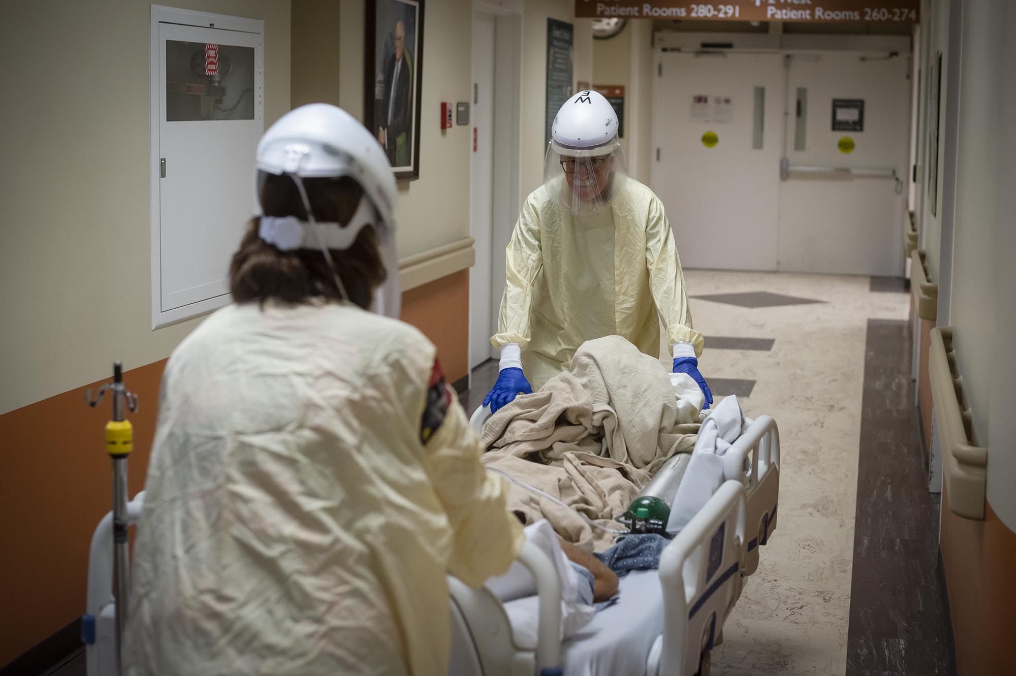 Two medical staff in protective gear push a patient on a hospital bed down a hallway. The patient is covered with a blanket. The hallway leads to a series of patient rooms.
