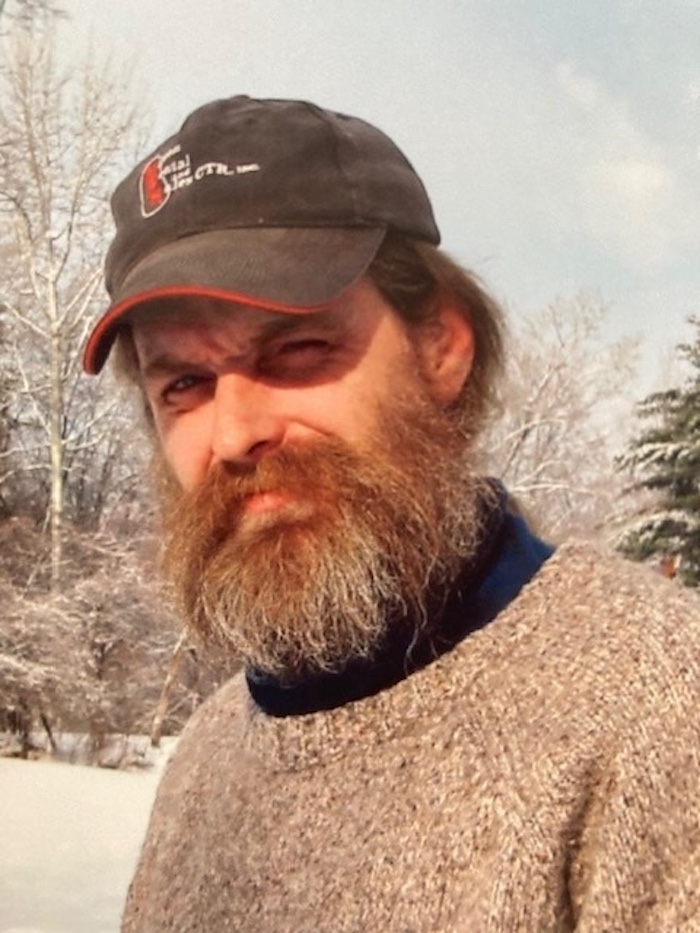 A man with a full beard wearing a knit sweater and a baseball cap stands outdoors on a snowy day with trees in the background.