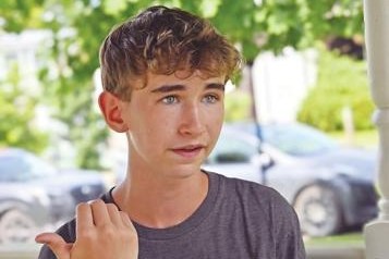 Teenager with short, wavy hair wearing a gray t-shirt gestures with his left hand while standing outdoors, with cars and greenery visible in the background.
