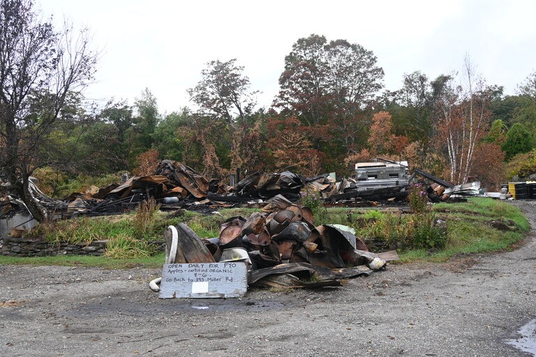 Debris and twisted metal from a burned-down structure lie scattered on the ground, with a sign in front and trees with autumn foliage in the background.