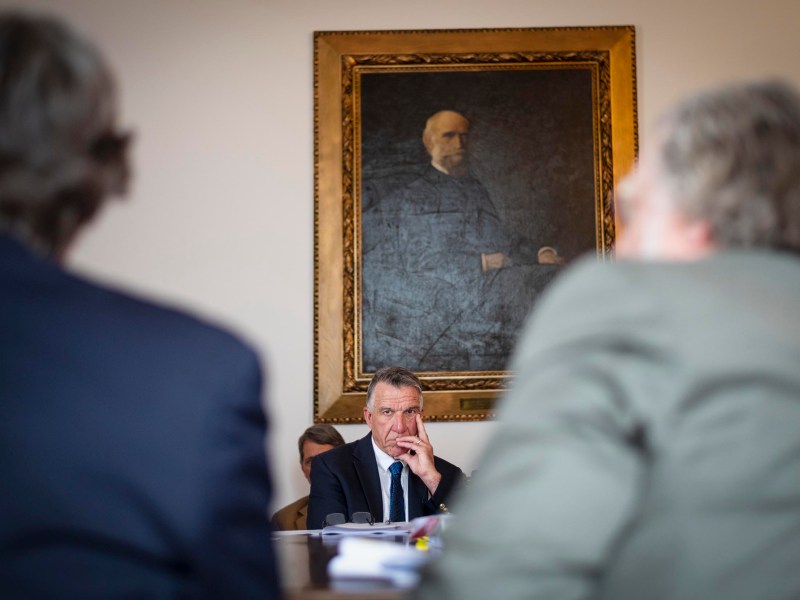 A man in a suit sits at a table, looking thoughtful, with a large framed portrait of an older man hanging on the wall behind him. Two blurred figures are in the foreground.