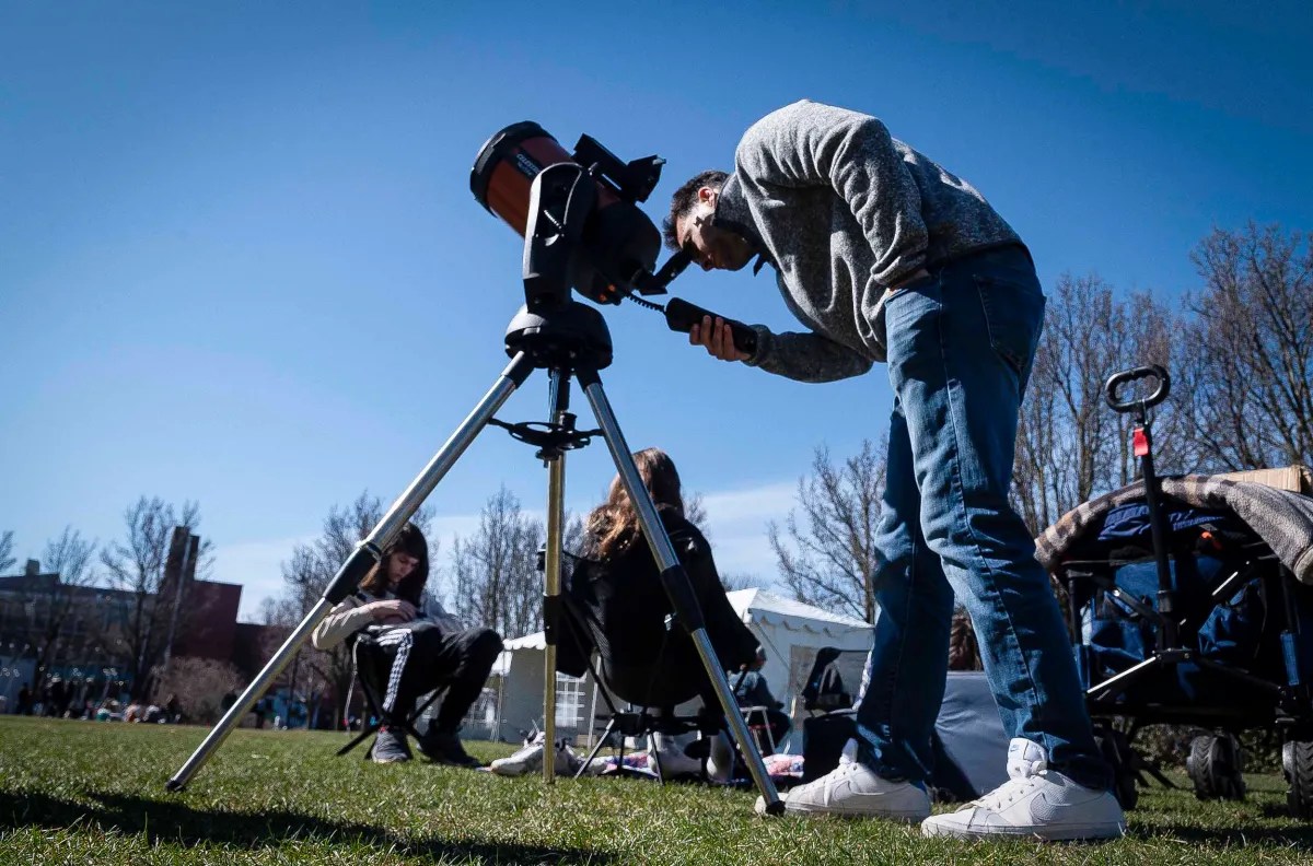 A person looking through a telescope during the daytime with others sitting on the grass nearby.