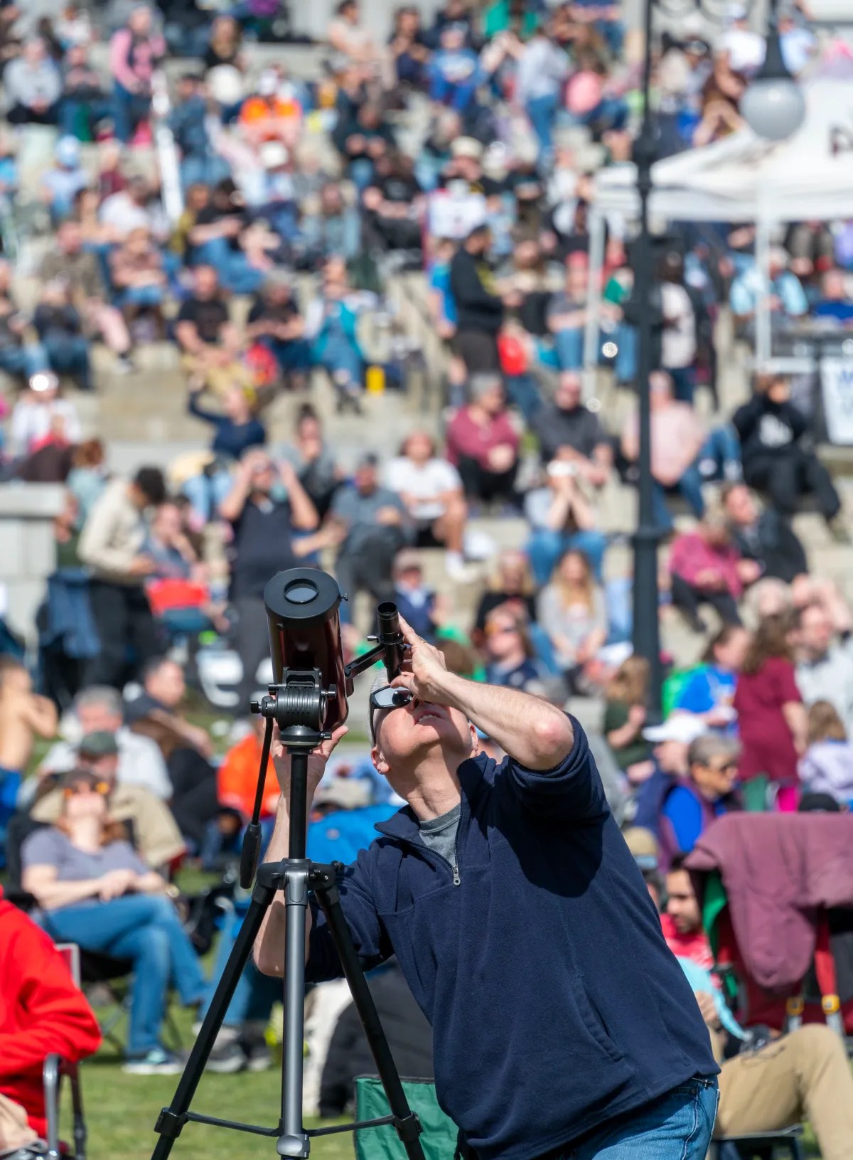 A man adjusts a telescope outdoors with a crowd of people seated in the background.