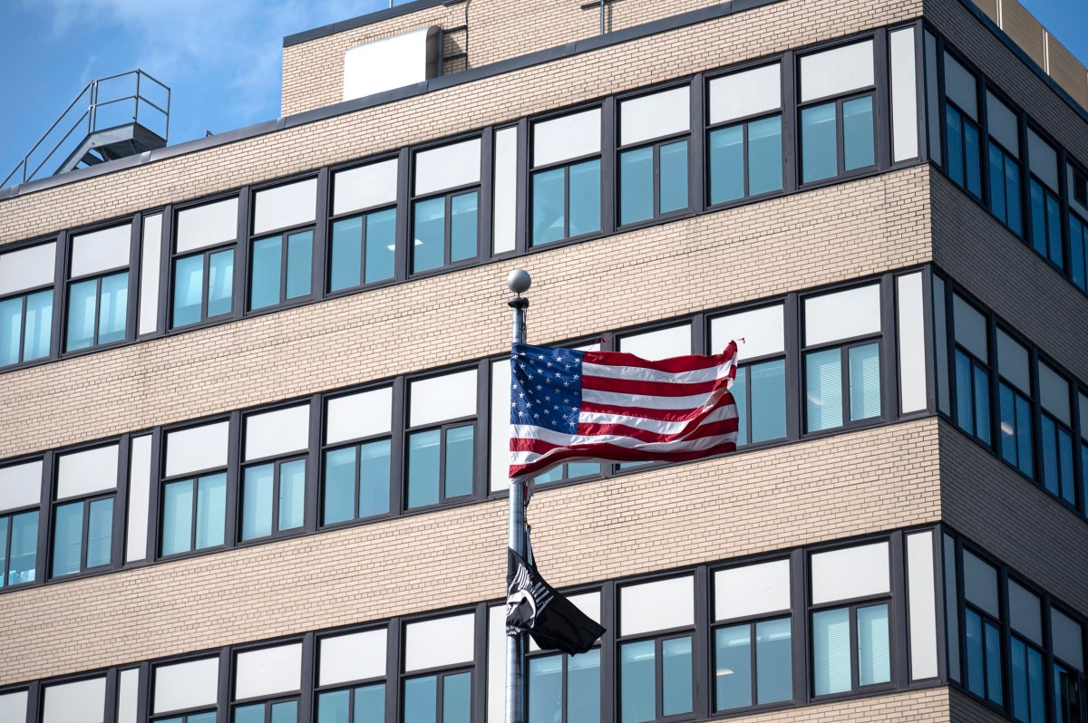 A U.S. flag and a black flag fly in front of a modern brick and glass office building under a clear sky.