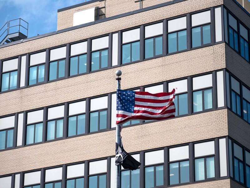 A U.S. flag and a black flag fly in front of a modern brick and glass office building under a clear sky.