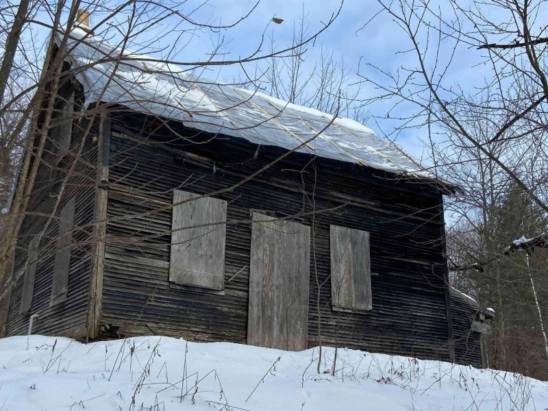 A weathered, wooden building with boarded windows and door stands on a snow-covered slope, surrounded by leafless trees under a cloudy sky.