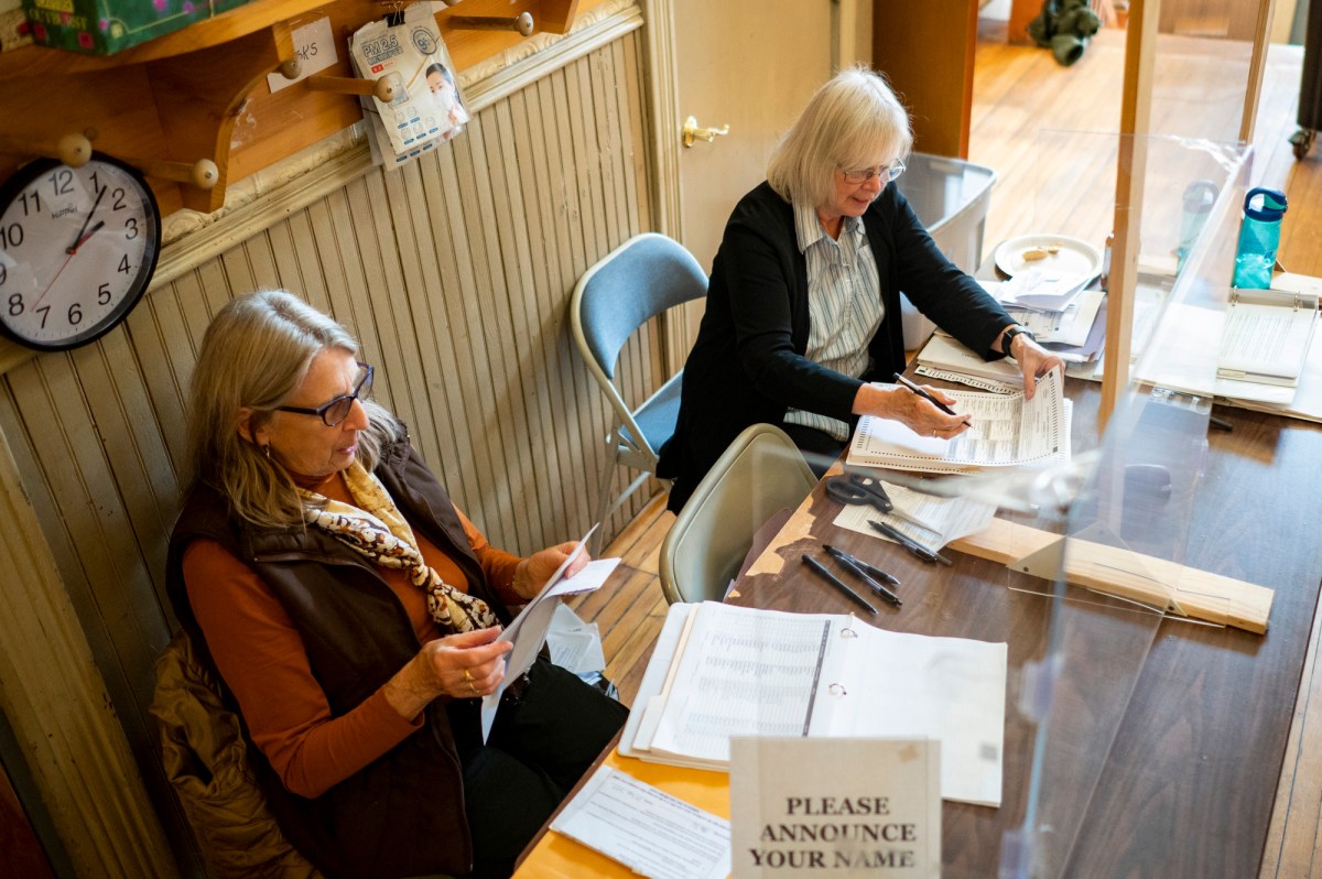 Two women sit at a table handling paperwork in an office. A sign reads, "Please announce your name.
