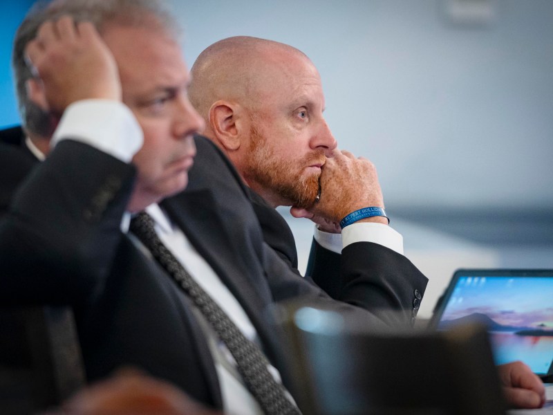 Two men in suits sit attentively at a meeting, with one man resting his chin on his hand in thought. Laptops are open in front of them.
