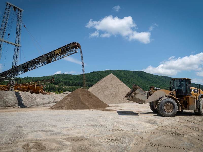 A large construction vehicle moves dirt next to a conveyor belt depositing material into a large pile at a quarry, with green hills and a blue sky in the background.