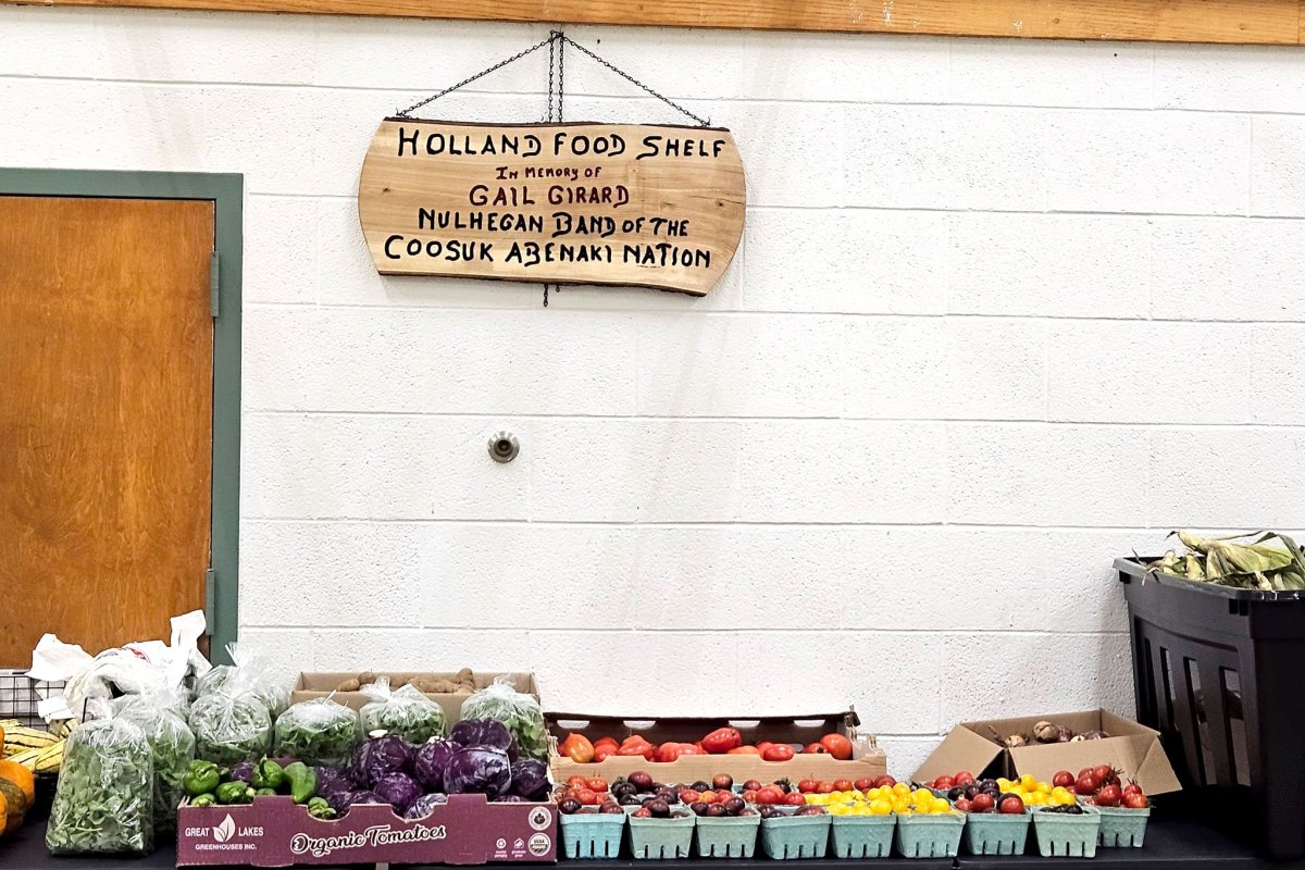 A table displays assorted fresh produce below a wooden sign reading “Holland Food Shelf in memory of Gail Girard, Nulhegan Band of the Coosuk Abenaki Nation.”.