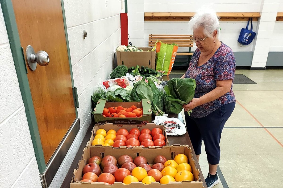 A woman selects leafy greens from a table displaying various fresh vegetables, including tomatoes, in a community center setting.