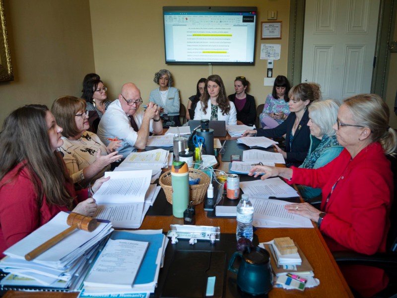 A group of people sit around a conference table covered with documents, notebooks, and drinks, holding a meeting with a screen displaying a document in the background.