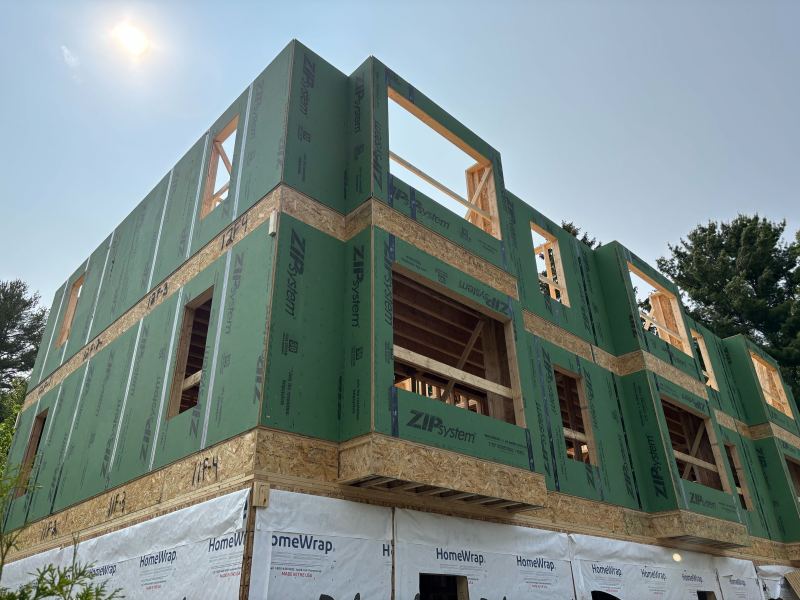 A partially constructed building with green ZIP System sheathing and exposed wooden framing under a sunny sky. The lower section is wrapped in white HomeWrap material.