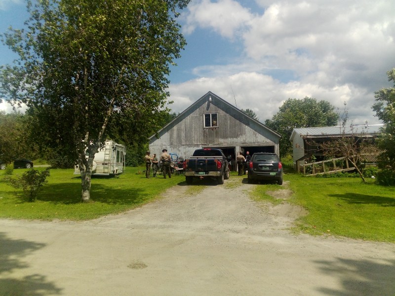 A group of people and vehicles gathered outside a large, weathered barn with an open garage on a sunny day.