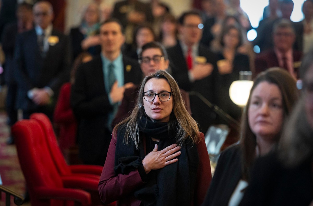 A group of people stands indoors with hands over their hearts. The setting appears formal with red seating.