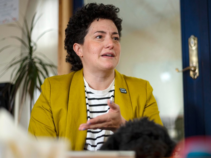A person wearing a yellow blazer and striped shirt gestures while speaking indoors, with a plant and door in the background.