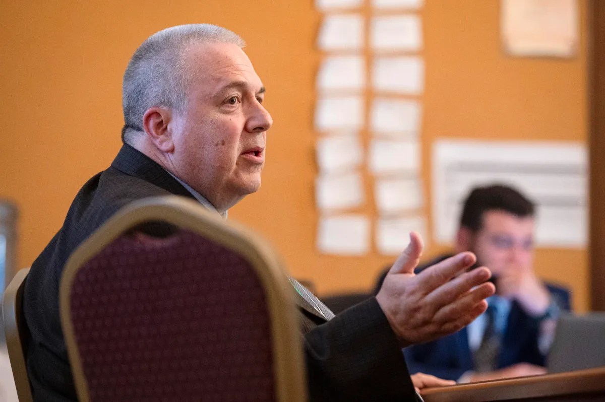 A man in a suit speaks while seated at a table in a meeting room; another person is visible in the background, blurred.