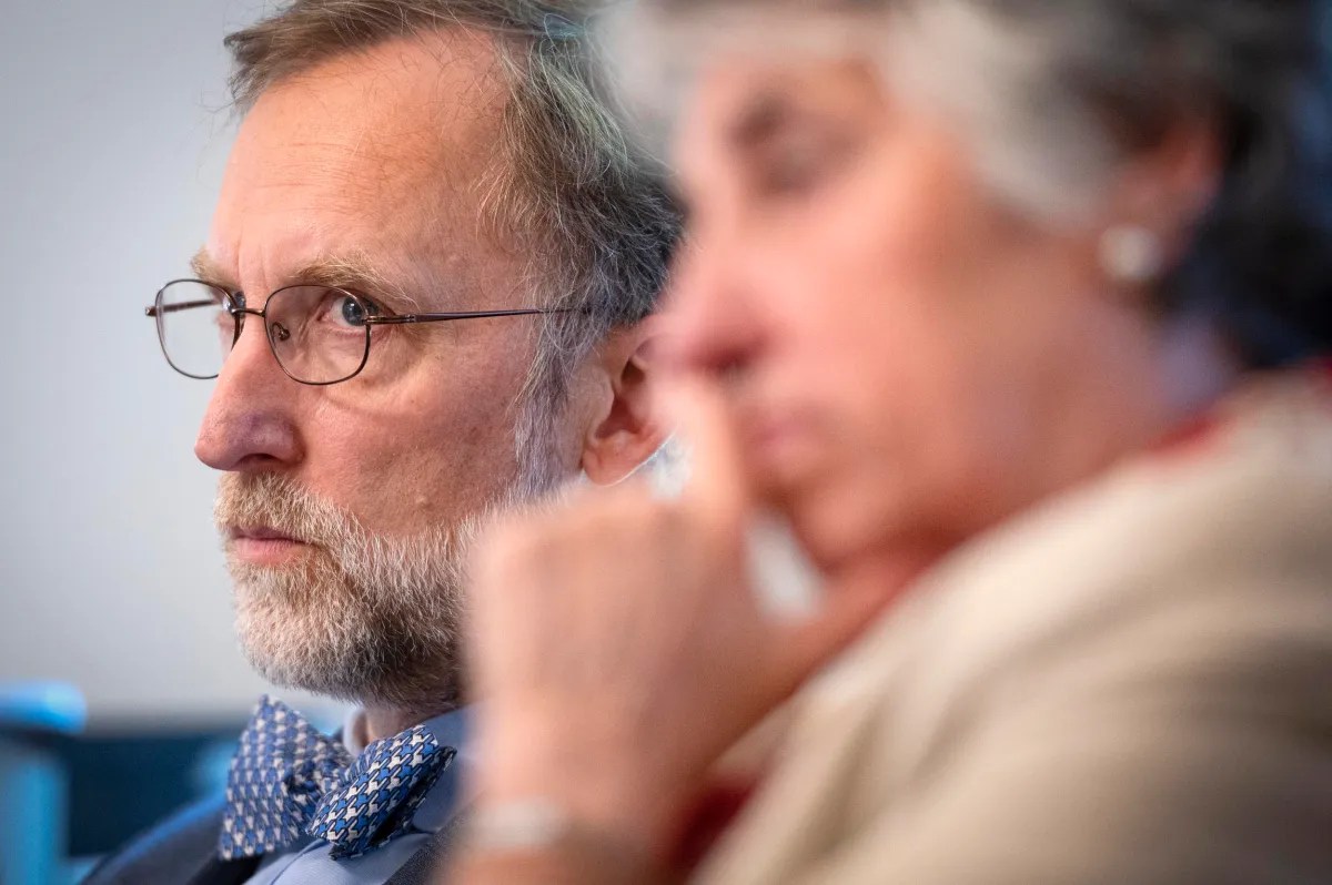 A man with glasses and a bow tie looks intently ahead while a woman in the foreground appears out of focus.