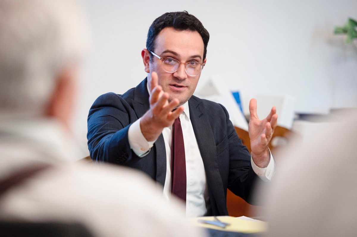A man in a suit and glasses gestures with his hands while speaking to others in a meeting setting.