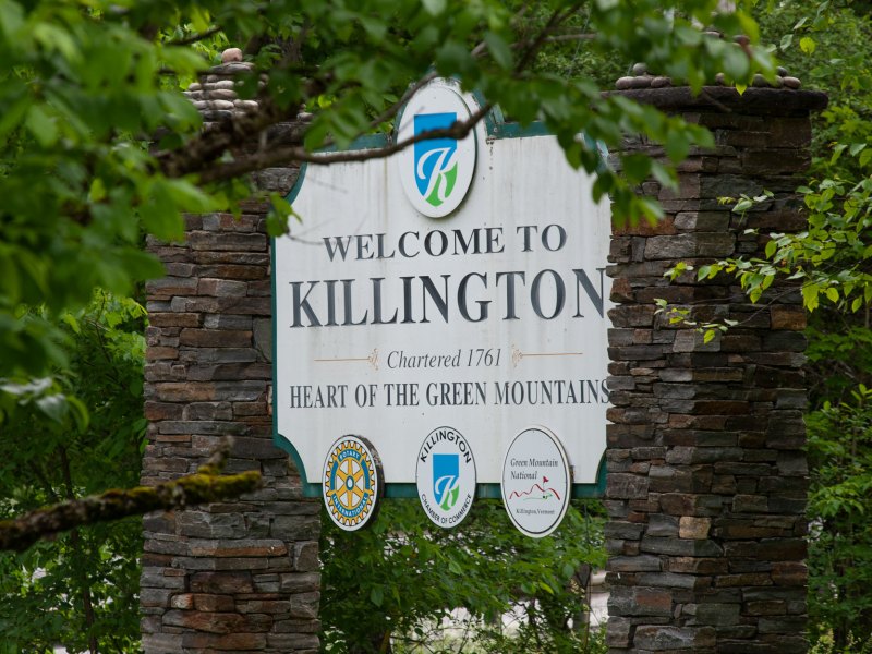 A stone sign reading "Welcome to Killington, Chartered 1761, Heart of the Green Mountains" is partially obscured by green foliage.