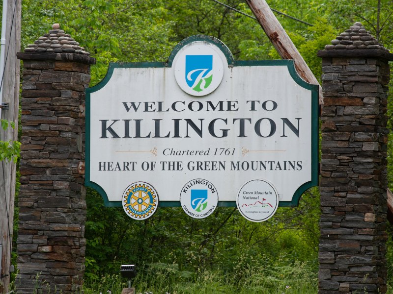Sign reading "Welcome to Killington, Chartered 1761, Heart of the Green Mountains," with logos of Rotary International, Killington sight, and Green Mountain National Forest, set against a background of trees.