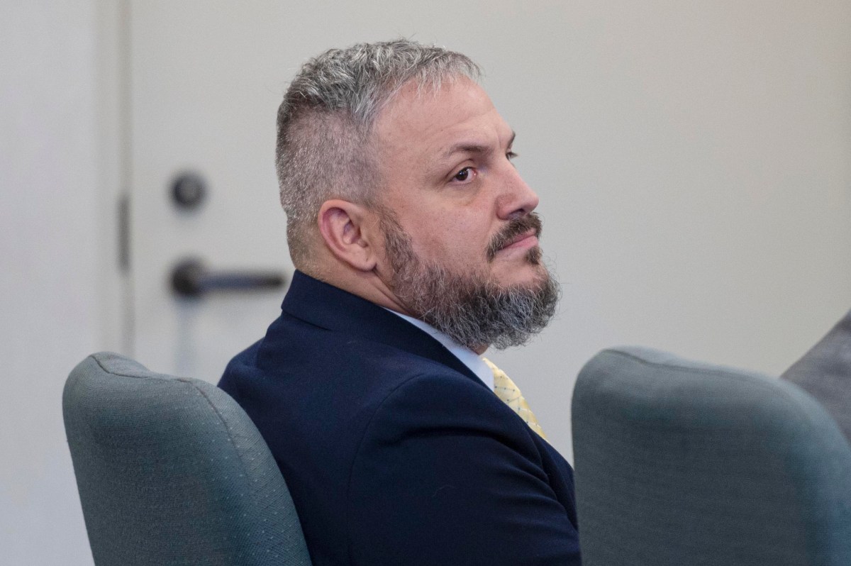 A man with short gray hair and a beard, dressed in a suit and yellow tie, sits in a chair facing slightly to the left in a room with plain walls.