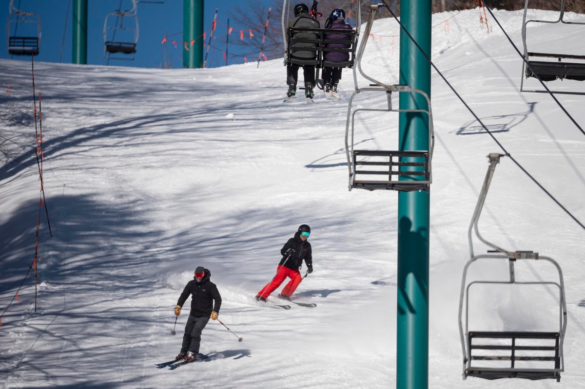 Two skiers in motion on a snowy slope under a blue ski lift carrying three people.