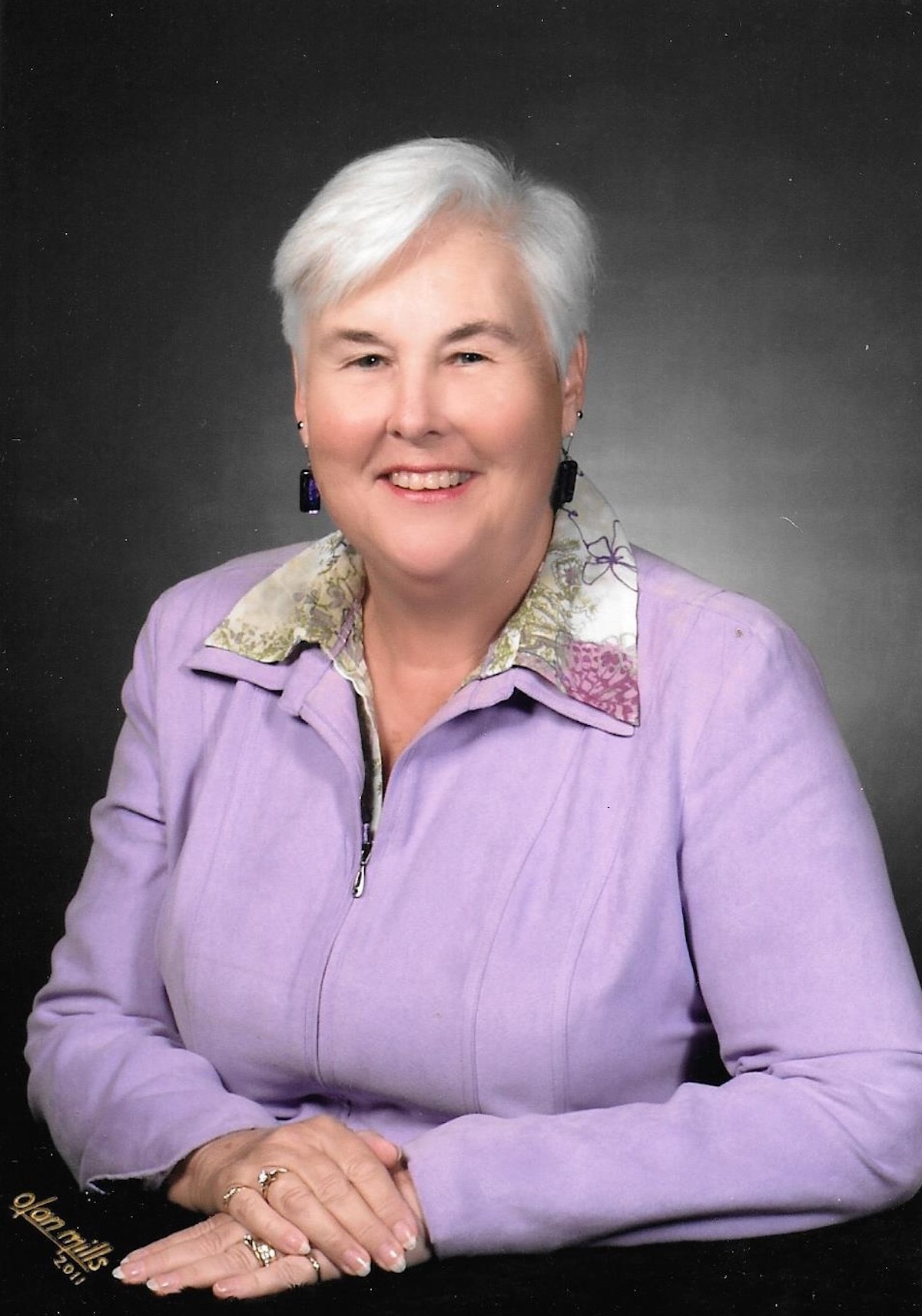 A woman with short hair wearing a light purple jacket poses for a formal studio portrait against a dark background.
