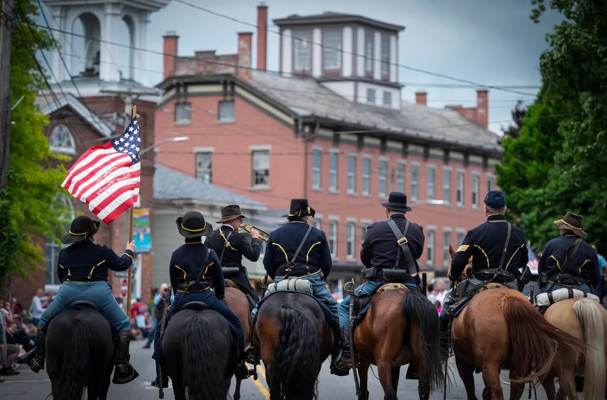 A group of six uniformed individuals on horseback ride in a parade, one holding an American flag, with buildings and spectators visible in the background.