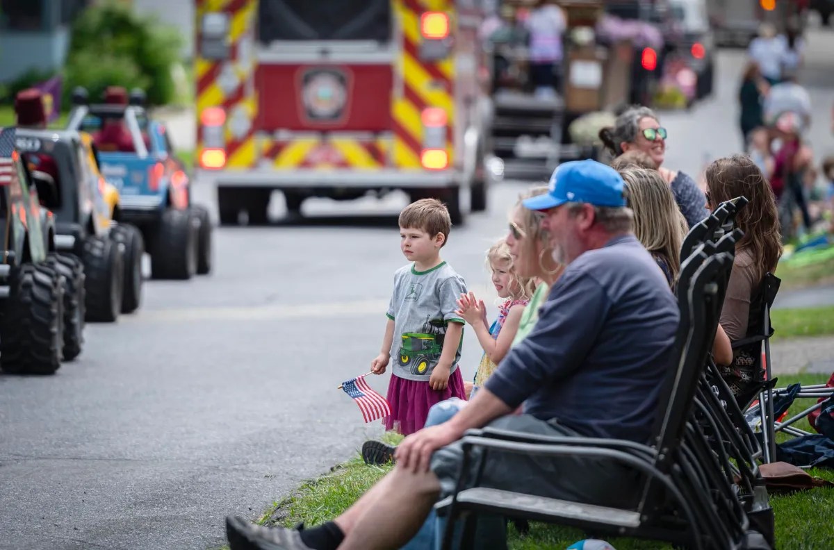 A group of people sitting and standing along a street, watching a parade with children holding American flags. A fire truck and miniature vehicles are visible in the background.
